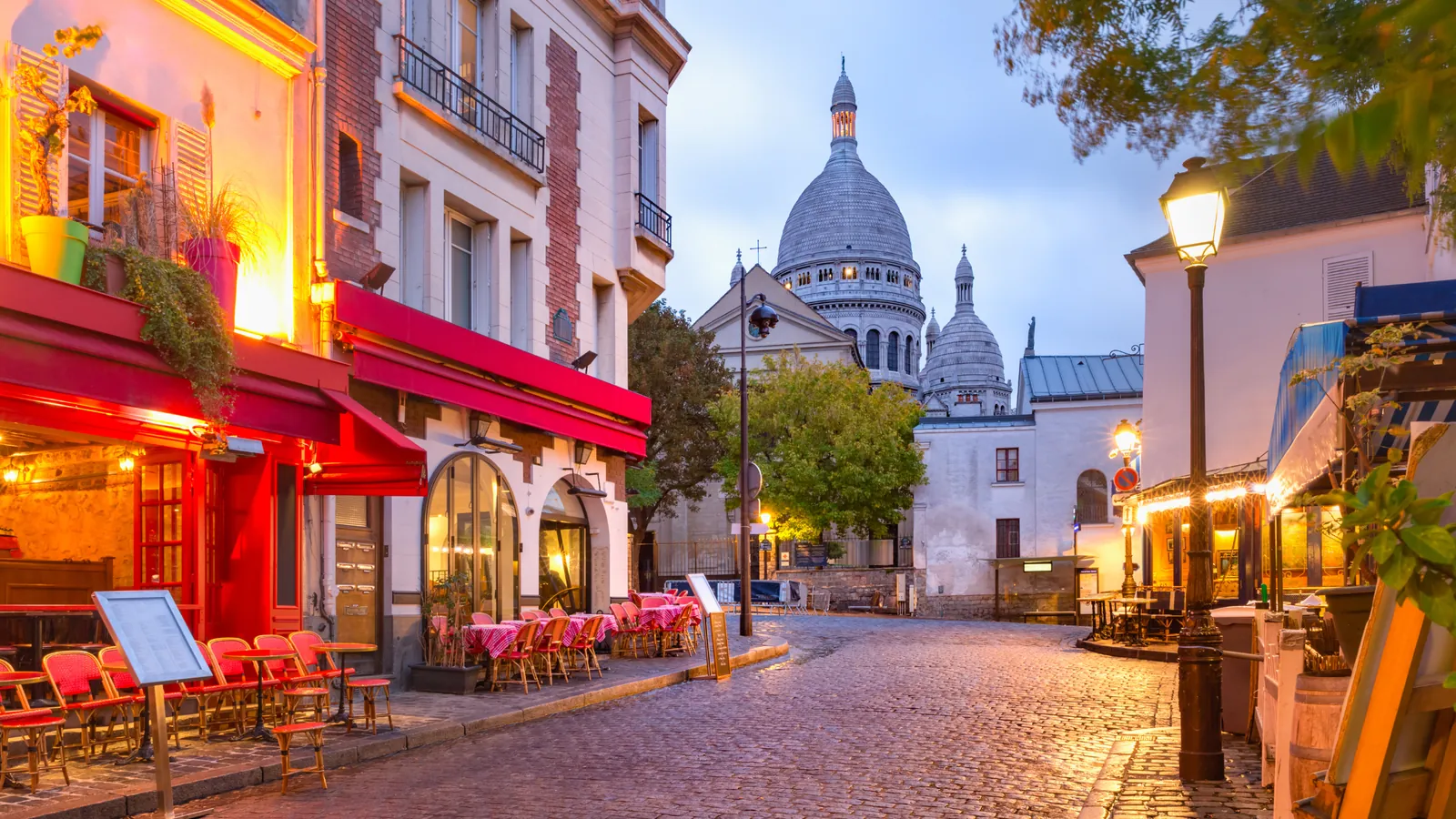 A café-lined cobbled street in Montmartre with the Sacré-Cœur basilica behind